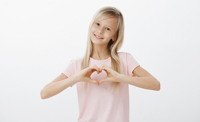 Cute trendy young girl loving her cat. Indoor shot of adorable little woman with pleased expression, showing heart gesture and smiling broadly, confessing in love to family over gray wall.